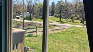 A room with a large window, a wooden chair, and a view of a grassy area with trees and a road. - Cozy 2-bedroom bungalow in gorgeous Mount Storm with AC (Mount Storm)