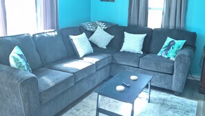 A living room with a sectional sofa, a coffee table, a ceiling fan, and a window with curtains. - Cozy 2-bedroom bungalow in gorgeous Mount Storm with AC (Mount Storm)