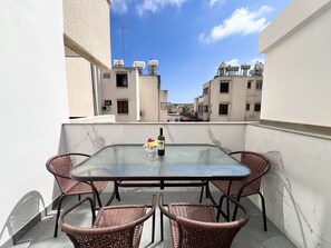 A glass-top table with wicker chairs on a balcony with a view of buildings. - 06 AN - Stay Monte Paradise (Ayia Napa)