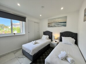 A hotel room with two beds, a window with a view, a painting on the wall, and a nightstand with a lamp. - 06 AN - Stay Monte Paradise (Ayia Napa)