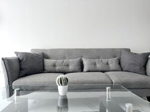 A grey sofa with multiple cushions, a glass coffee table with a cactus, and a white vase. - 06 AN - Stay Monte Paradise (Ayia Napa)