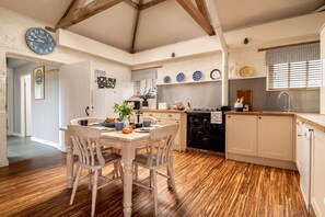 Une cuisine avec un plancher en bois, une table à manger avec des chaises, une horloge et une fenêtre avec des stores.