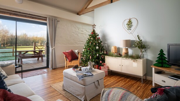 Una sala de estar acogedora con piso de madera, un sofá blanco, un pequeño reposapiés, un árbol de Navidad y un televisor sobre un mueble de madera.