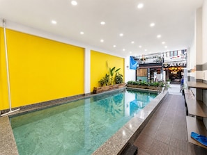 An indoor swimming pool with a yellow wall and a restaurant in the background. - Two Horses Hostel (Hoi An)