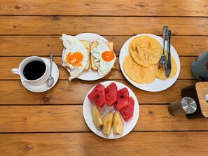 Une table en bois sur laquelle sont disposés des aliments pour le petit-déjeuner, notamment des toasts avec des œufs, des crêpes, une tasse de café, des tranches de pastèque et des tranches de pomme.