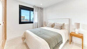 A modern bedroom with a large bed, a nightstand, a window with curtains, and a city view. - LXR Dalí 1-Bedroom Beachfront (Fuengirola)