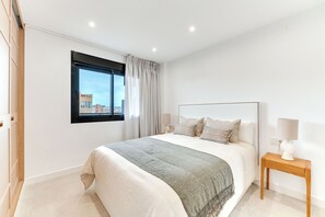 A modern bedroom with a large bed, a nightstand, a window with curtains, and a city view. - LXR Dalí 1-Bedroom Beachfront (Fuengirola)