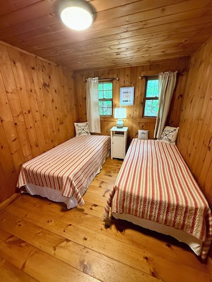 Ein blockhausspezifisches Zimmer mit zwei Betten, Holzdielen, einem Nachttisch mit Lampe und einem fenster mit Vorhängen.