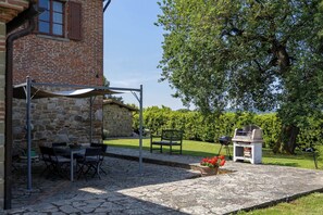 Un patio con pavimento in pietra, un tavolo con sedie, un barbecue e un grande albero che offre ombra.