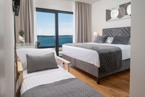 Ein Hotelzimmer mit zwei Betten, einem großen Fenster mit Meerblick, einem Nachttisch mit Lampe und einem an der Wand hängenden Kunstwerk.