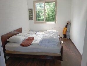 Una cama individual con estructura de madera, una ventana que da a árboles, y un cuadro colgado en la pared.