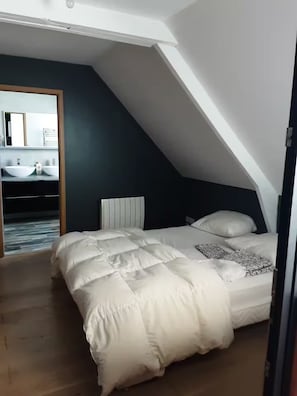 Quarto com teto inclinado, cama com roupa de cama branca, cabeceira e um espelho grande sobre uma toucador com duas pias.