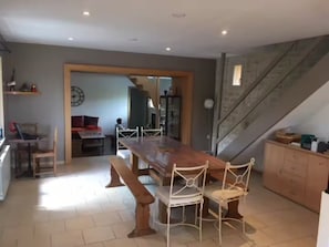 Uma ampla área de refeições com uma longa mesa de madeira e cadeiras de metal branco, uma cozinha com armários e uma escada.