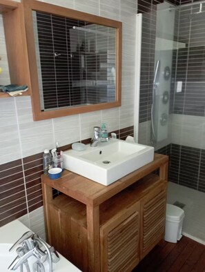 Un baño con tocador de madera, lavamanos blanco, espejo, cabina de ducha y pared con azulejos.