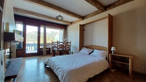 A bedroom with a large bed, a wooden headboard, a bedside table with a lamp, and a view of a balcony with a table and chairs. - Cosy studio with terrace (Gréolières)