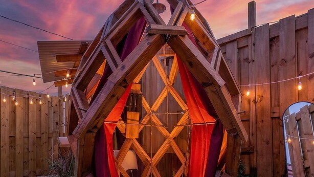 Een houten constructie met een ruitpatroonraam en rode gordijnen, omgeven door een grindgebied en een houten hek.