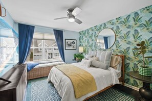 Une chambre avec un lit, un ventilateur de plafond, une fenêtre avec des rideaux, un mur orné d’un motif de feuilles tropicales et un miroir rond.