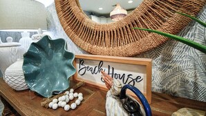 Ein Holztisch mit einem Schild mit der Aufschrift 'Beach House', dekorativen Gegenständen und einer gewebten Wandbehängung.