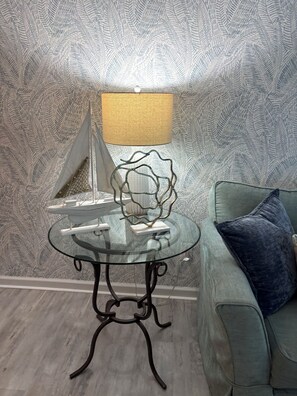 A room with a glass table, a lamp, a decorative item, a sailboat model, and a couch with a pillow.