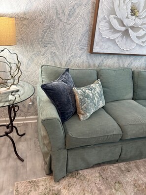 A green sofa with patterned cushions, a glass table with a metal base, and a large floral artwork on the wall.