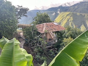 A rustic house with a tiled roof, surrounded by lush greenery and a mountainous backdrop.