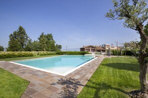 A rectangular swimming pool with a tiled deck, surrounded by well-maintained grass and trees, with a house in the background. - Apartment Delphine with shared Pool (Castelnuovo del Garda)