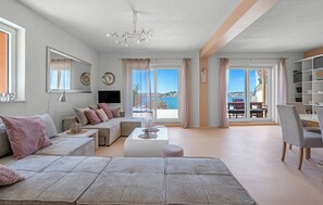 Ein geräumiges Wohnzimmer mit einem großen grauen Sofa, einem Couchtisch und einem Fernseher. Zwei große Fenster mit Vorhängen, eine Chandelüster und ein Bücherregal sind ebenfalls vorhanden.