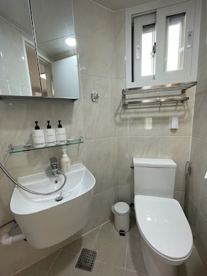 Un baño con lavamanos, inodoro y espejo blancos. Hay artículos de aseo en el lavamanos, y se ve una zona de ducha.