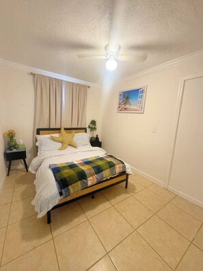 Ein Schlafzimmer mit Bett, Deckenventilator, Nachttisch, Fenster mit Vorhängen und einem Wandbild.