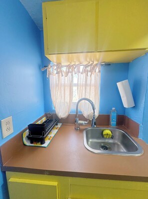 Eine kleine Küche mit Spüle, Wasserhahn und einem Fenster mit durchsichtigen Vorhängen.
