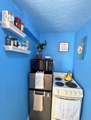 Eine Küche mit blauer Wand, einer Mikrowelle, einem Herd und einem Kühlschrank. Es gibt Regale mit Küchenutensilien und eine Pflanze.