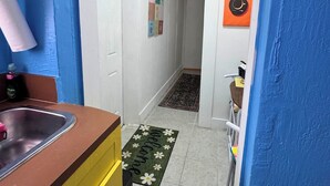 A narrow hallway with a kitchen area on one side, a door, a wall-mounted clock, and a small rug on the floor. - 1 bedroom Private Studio with Patio. 5 Min to Hard Rock Stadium & Every Event. (Miami Gardens)