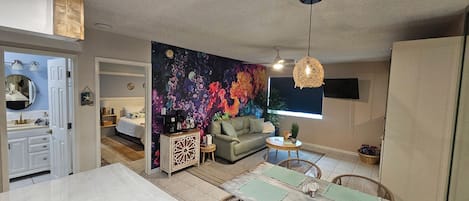 A living room with a colorful abstract mural, a sofa, a coffee table, and a dining area with chairs.