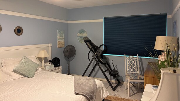 A bedroom with a bed, a ceiling fan, a standing fan, a treadmill, a window with blinds, and a bedside table with a lamp. - Chic & Comfortable Beach Home • Walk/Bike to the Coast • All Essentials Included (Pompano Beach)