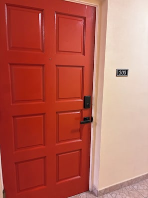 Eine rote Tür mit einem Zifferncode-Schloss, einem Klingelknopf und einem Schild mit der Zimmernummer.