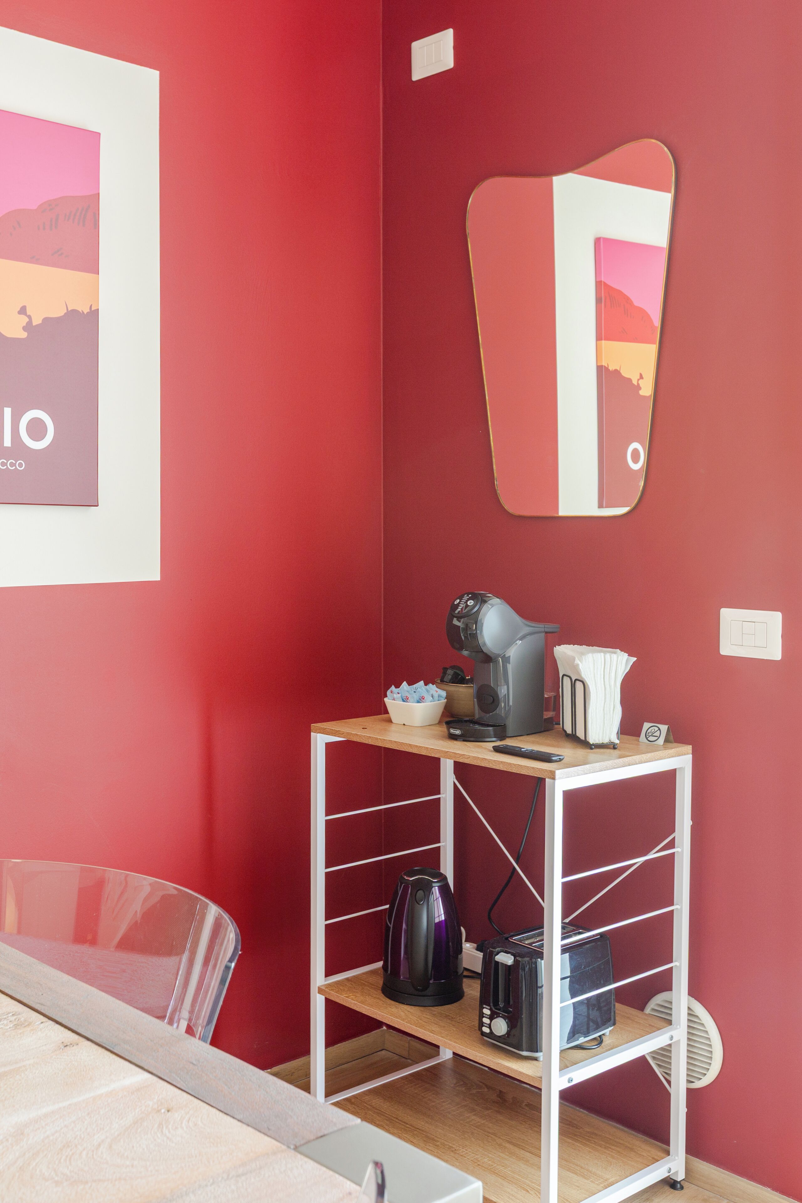 Une pièce avec un mur rouge, un miroir à cadre blanc, une étagère en bois sur laquelle se trouvent une bouilloire et un grille-pain, ainsi qu’une chaise transparente.