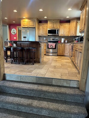 A kitchen with wooden cabinets, a stainless steel refrigerator, a microwave, a stove, and a bar area with stools.