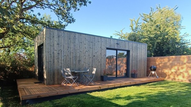Una estructura moderna de madera con puerta corredera de vidrio, un pequeño patio con mesa y sillas, y una parrilla.
