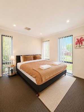 A modern bedroom with a large bed, a bedside table with a lamp, and a view through the window. - Modern Oasis | 3BR Home (Bassendean)
