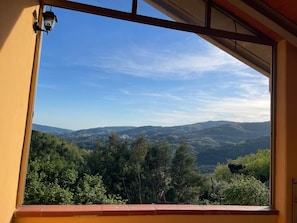A view from a window with a yellow wall and a metal frame, overlooking a landscape with hills and trees.