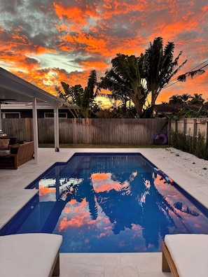 A swimming pool with a wooden deck, a covered patio area, and a view of a sunset with clouds. - Luxury beach house, heated pool, 10 minute walk to beach! (Pompano Beach)
