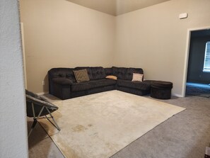 Ein Wohnzimmer mit Ecksofa, einem kleinen Hocker, einem weißen Teppich und einer Treppe, die zu einer weiteren Ebene führt.