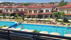 A pool area with lounge chairs and umbrellas, surrounded by buildings with red-tiled roofs. - Elegant Pool View 2 bedroom Apartment in Green Life Resort, Sozopol (Sozopol)