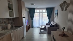 A modern living room with a sofa, a coffee table, and a TV. There is a kitchen area with cabinets and a sink. - Elegant Pool View 2 bedroom Apartment in Green Life Resort, Sozopol (Sozopol)