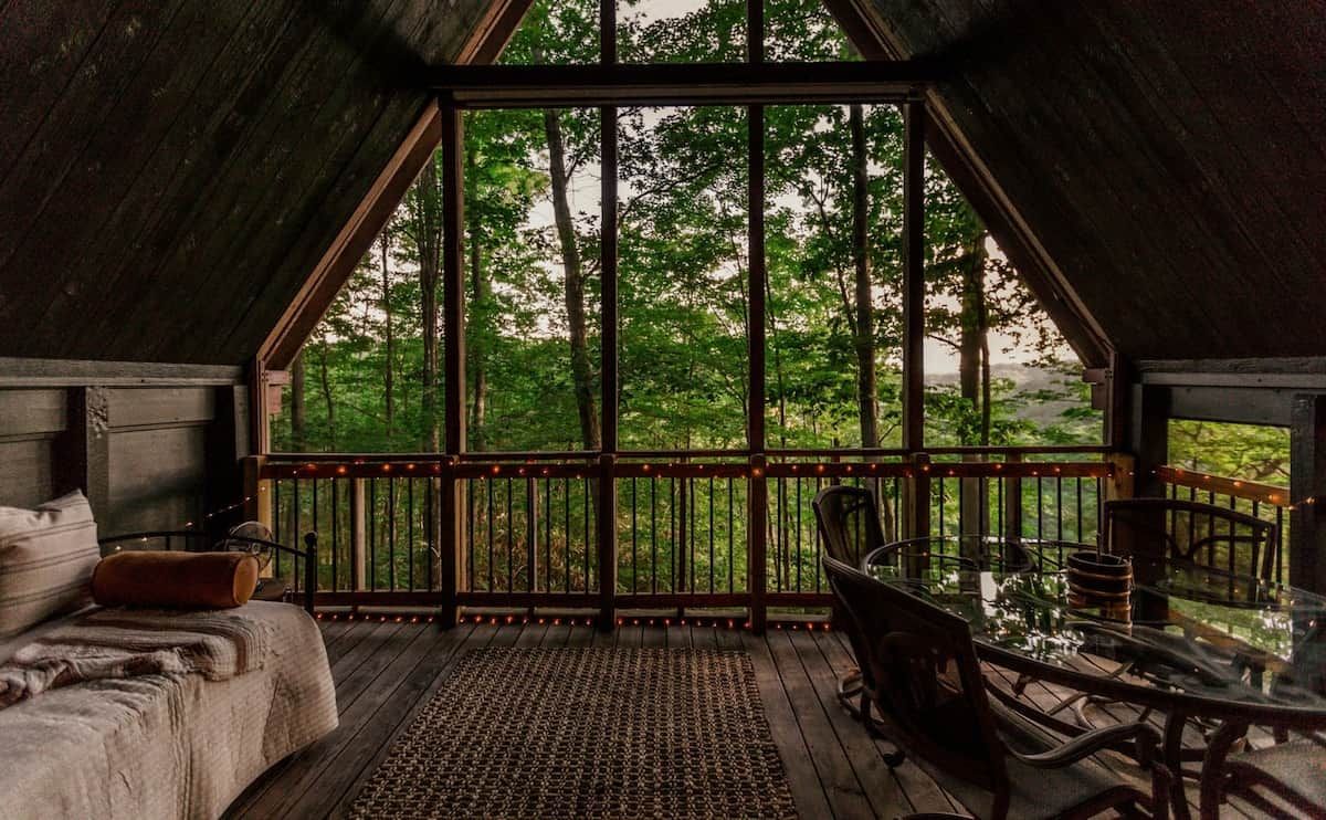 A cozy cabin with a bed, a dining table, and a view of a forest through large windows.