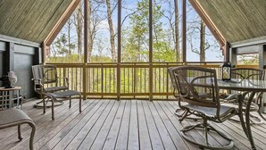 A covered outdoor area with a table and chairs, a view of trees through the railing, and a wooden floor.
