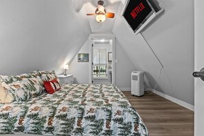 Ein Schlafzimmer mit einem Bett, einem Deckenventilator, einem an der Wand befestigten Fernseher, der 'NETFLIX' zeigt, und einem Fenster mit Jalousien.
