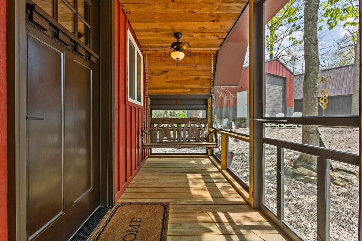 A wooden porch with a bench, a door, and a view of a red building and trees.