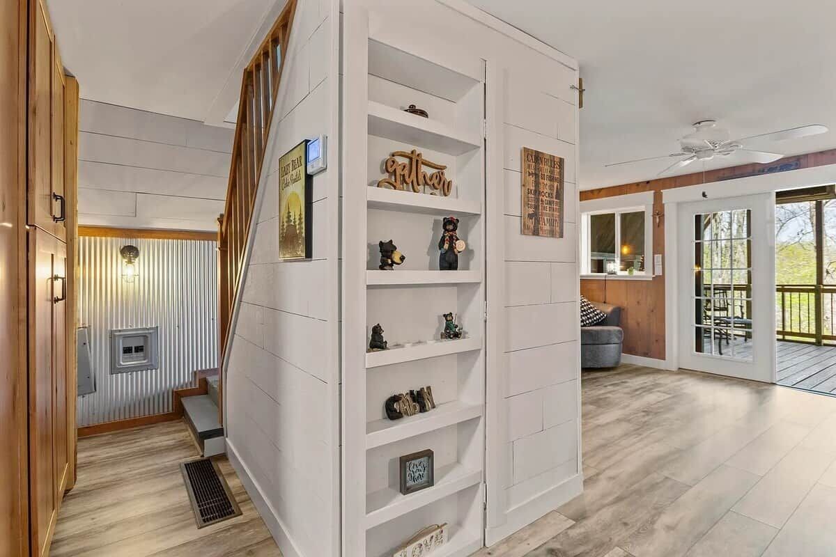 A modern interior with a staircase, white shelving with decorative items, and a view to an outdoor patio area.