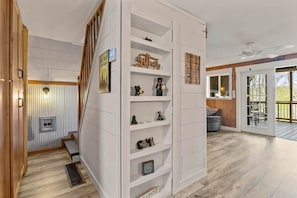 A modern interior with a staircase, white shelving with decorative items, and a view to an outdoor patio area.